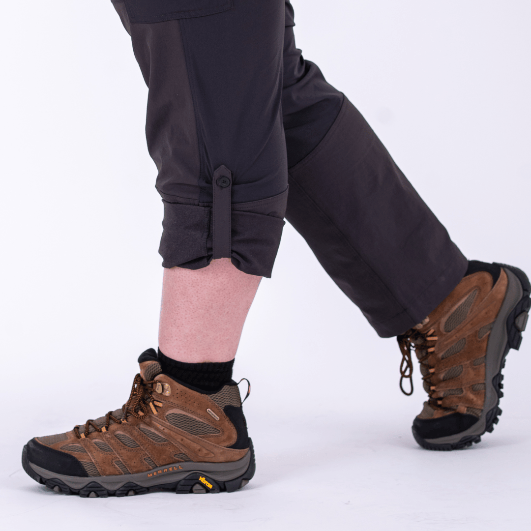 A pair of gray pants with roll tabs holding up one of the legs on a person with hiking boots.