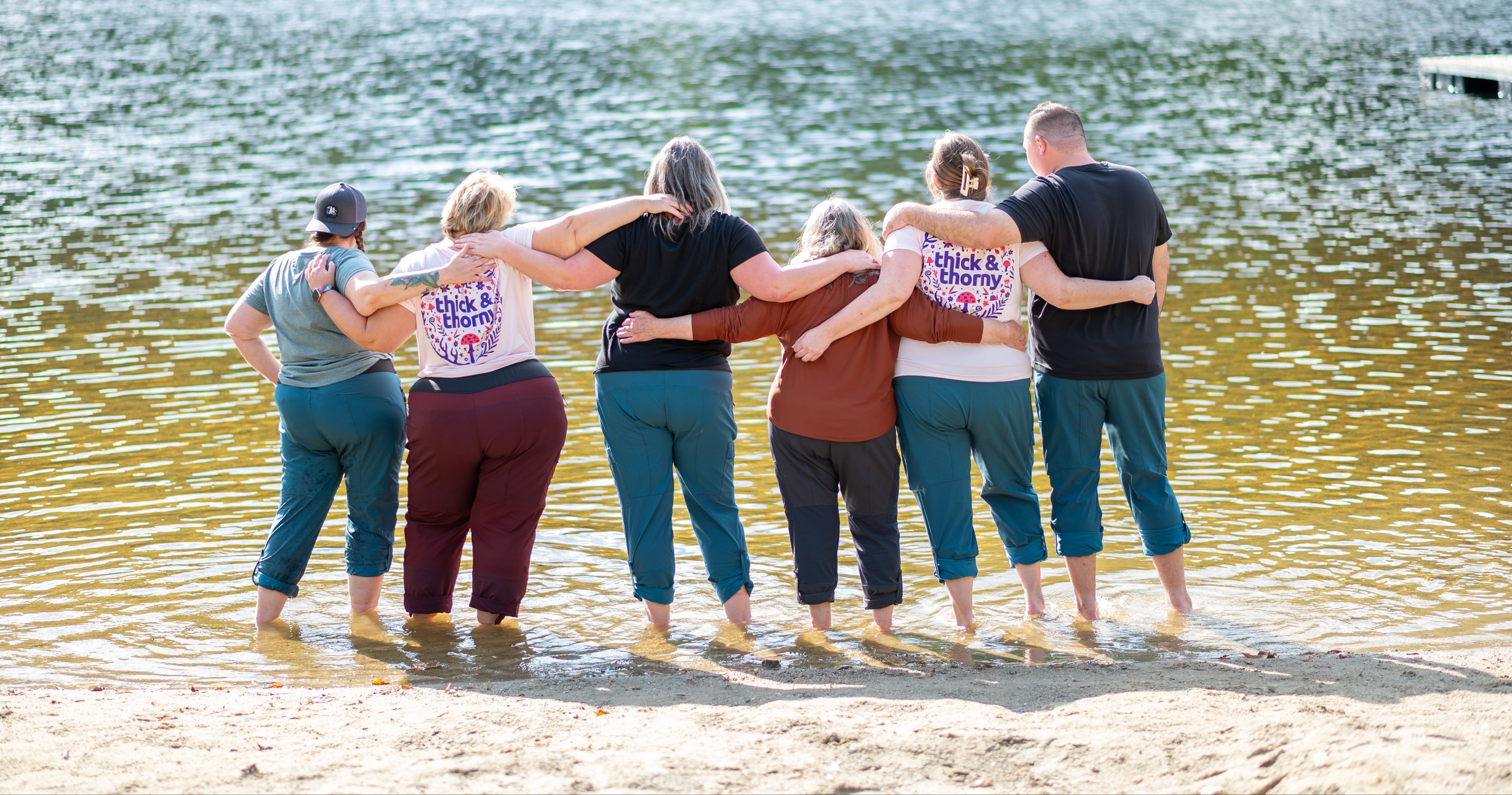 Group of people standing in a line with arms around each other on a lakeside.