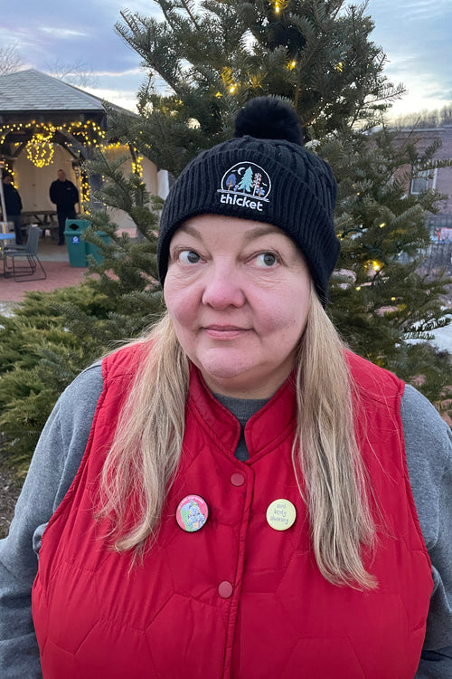 Person wearing a red vest and black beanie with a logo, standing in front of a decorated Christmas tree.