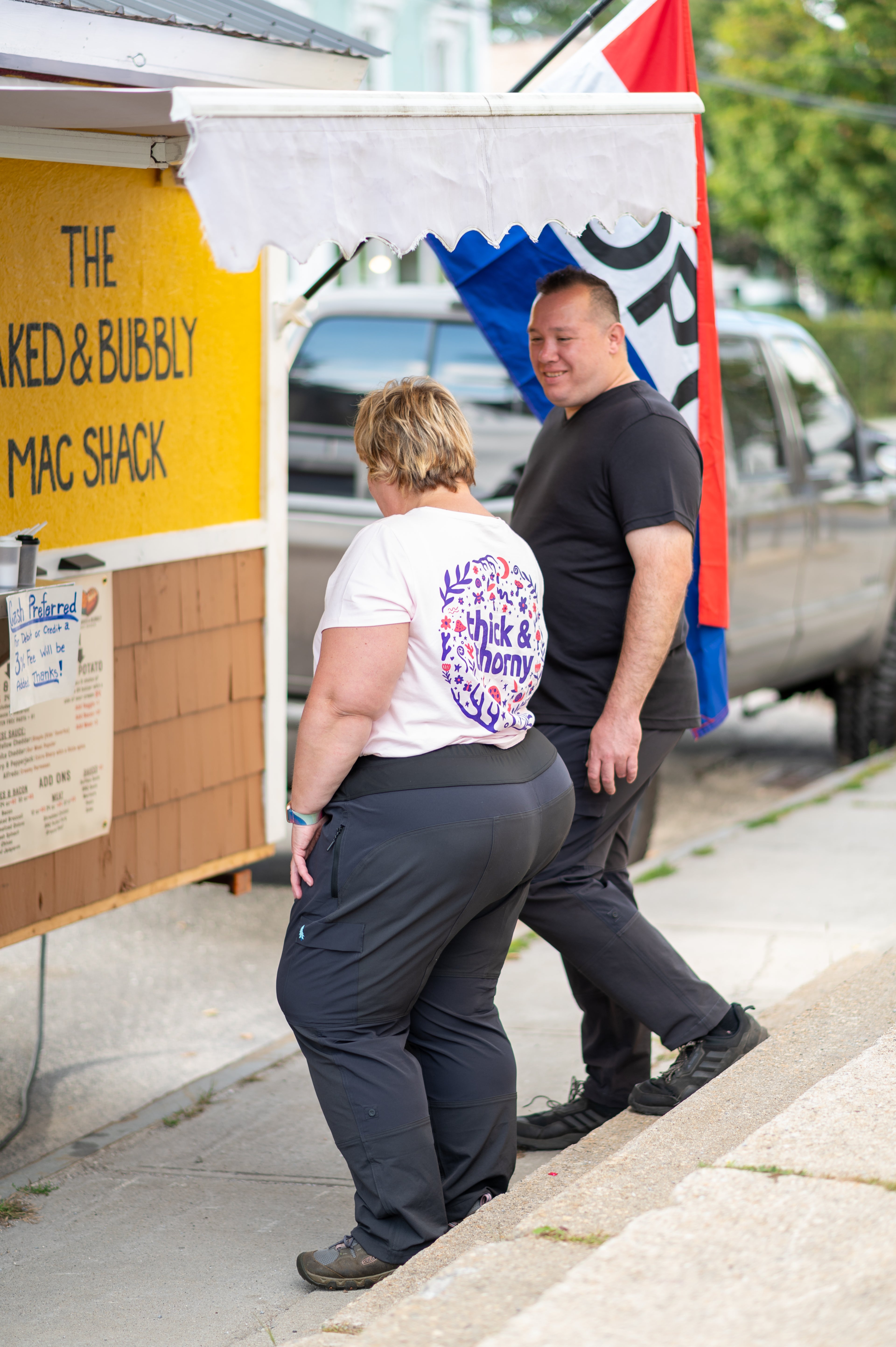 Two people standing next to a food stand with a sign reading 'The Naked & Bubbly Mac Shack'.